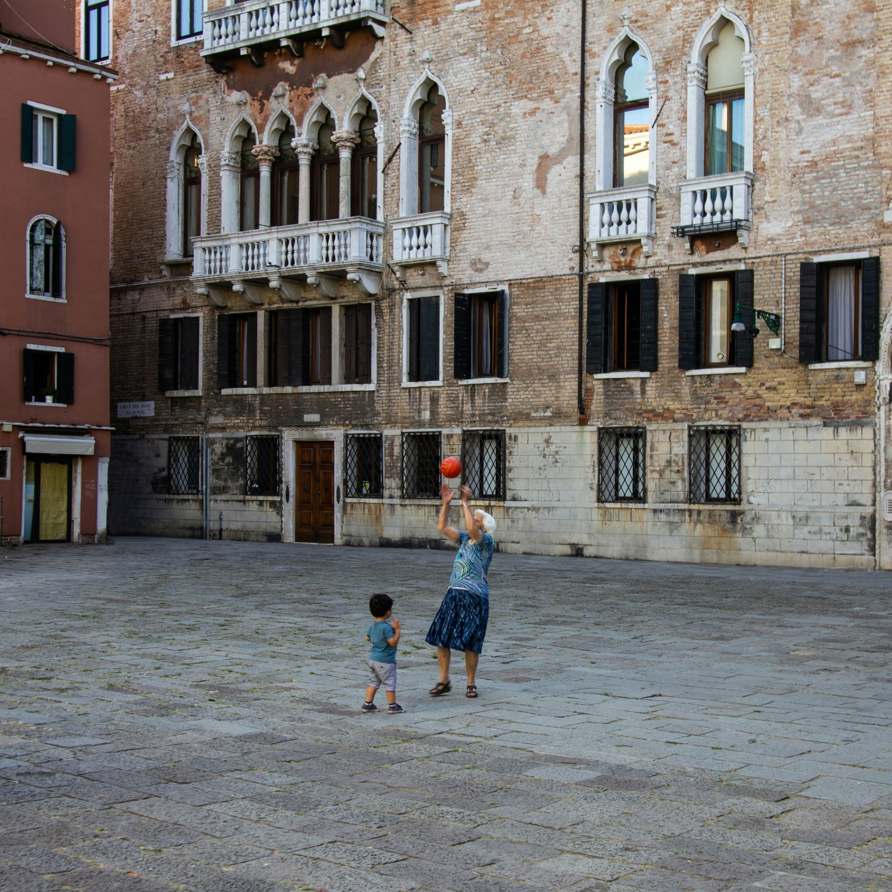 An elderly woman and a child playing with a basketball in a Venetian piazza.