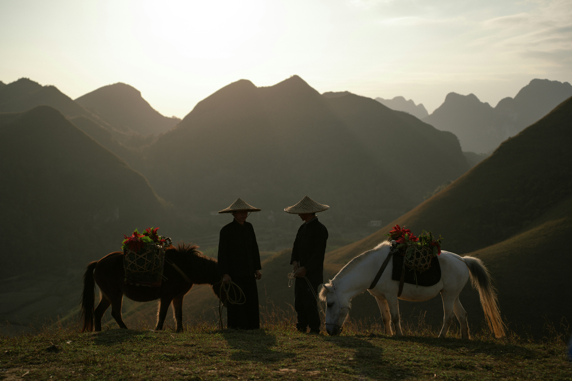 Two figures in conical hats stand with their horses on a high ridge as the sun sets behind layered mountains.